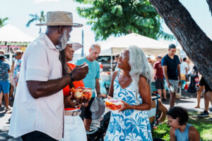 Vibrant senior woman holding fruit talking to an African American couple at a farmers market on a sunny day.